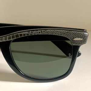May include: Black sunglasses with a textured, patterned top frame and dark green lenses. The sunglasses have a classic, angular shape and a small silver accent on the frame.