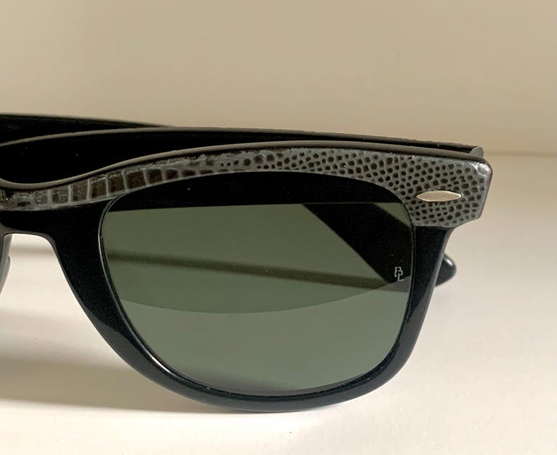 May include: Black sunglasses with a textured, patterned top frame and dark green lenses. The sunglasses have a classic, angular shape and a small silver accent on the frame.