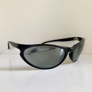 May include: Black wrap-around sunglasses with dark grey lenses. The frames feature a smooth, curved design with the Ray-Ban logo printed in white on the upper corner of the right lens. The temples are thin and connect to the front frame with hinges.