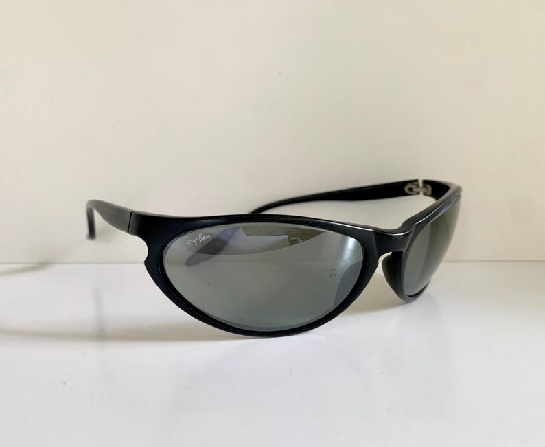 May include: Black wrap-around sunglasses with dark grey lenses. The frames feature a smooth, curved design with the Ray-Ban logo printed in white on the upper corner of the right lens. The temples are thin and connect to the front frame with hinges.