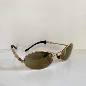 May include: Gold-toned sunglasses with oval lenses and black temples. The sunglasses have a sleek, modern design with a subtle curve to the frame. The lenses are a brown tint, providing a contrast to the gold frame.