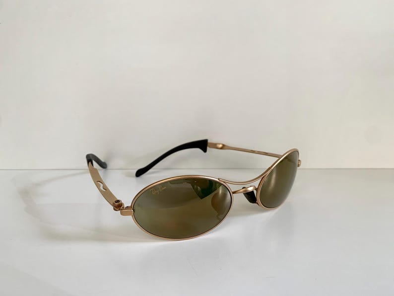 May include: Gold-toned sunglasses with oval lenses and black temples. The sunglasses have a sleek, modern design with a subtle curve to the frame. The lenses are a brown tint, providing a contrast to the gold frame.