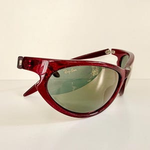 May include: A pair of Ray-Ban sunglasses with a deep red frame and green lenses. The sunglasses have a unique, curved design and the Ray-Ban logo is visible on the lens. The frame has a glittery texture.