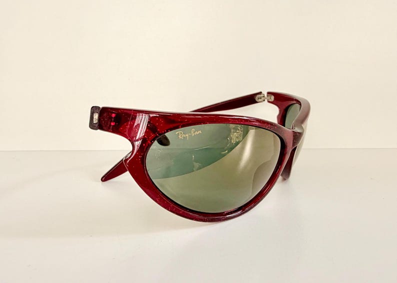 May include: A pair of Ray-Ban sunglasses with a deep red frame and green lenses. The sunglasses have a unique, curved design and the Ray-Ban logo is visible on the lens. The frame has a glittery texture.