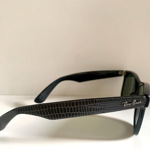 May include: Black sunglasses with a textured, dark gray pattern on the arms. The Ray-Ban logo is visible on the side. The lenses are dark, and the frames are black.