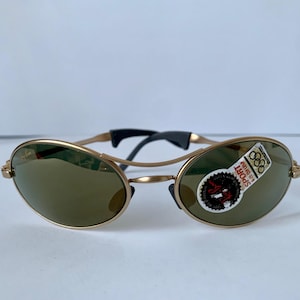 May include: Gold-tone sunglasses with oval lenses and a black bridge. The sunglasses have a sticker on the lens with the Olympic rings and the word "SPORT". The frames are a light gold color.