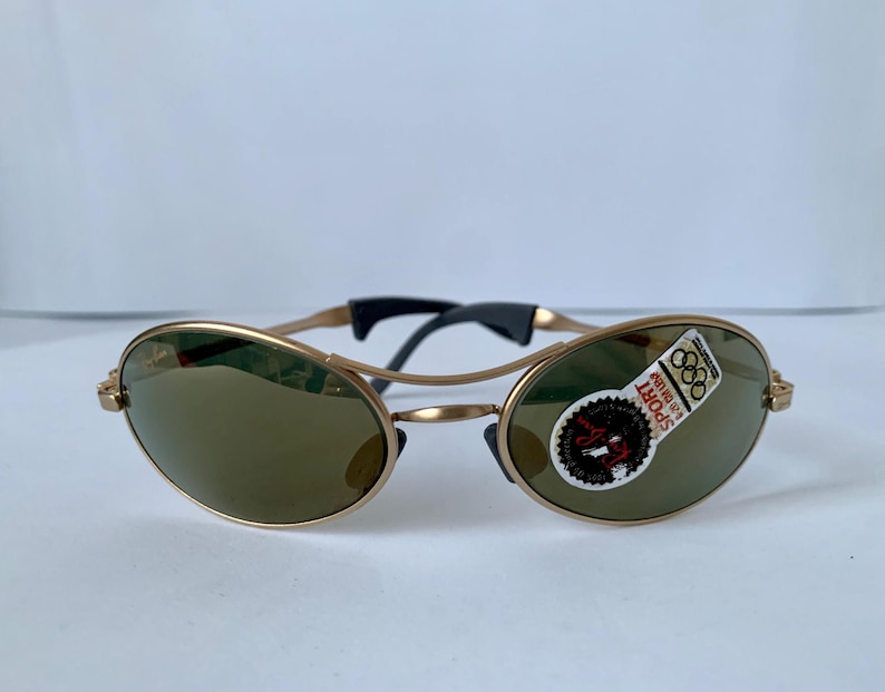 May include: Gold-tone sunglasses with oval lenses and a black bridge. The sunglasses have a sticker on the lens with the Olympic rings and the word "SPORT". The frames are a light gold color.