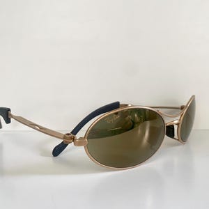 May include: Gold-tone Ray-Ban sunglasses with oval lenses. The sunglasses have a gold frame, brown lenses, and black accents on the temples. The Ray-Ban logo is visible on the lens.