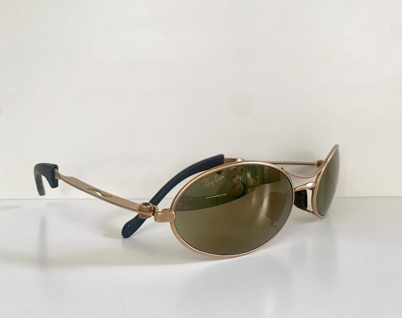 May include: Gold-tone Ray-Ban sunglasses with oval lenses. The sunglasses have a gold frame, brown lenses, and black accents on the temples. The Ray-Ban logo is visible on the lens.