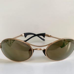 May include: Oval-shaped sunglasses with a gold-colored frame and dark lenses. The sunglasses have a double bridge design and black temples. The lenses reflect the surrounding environment.