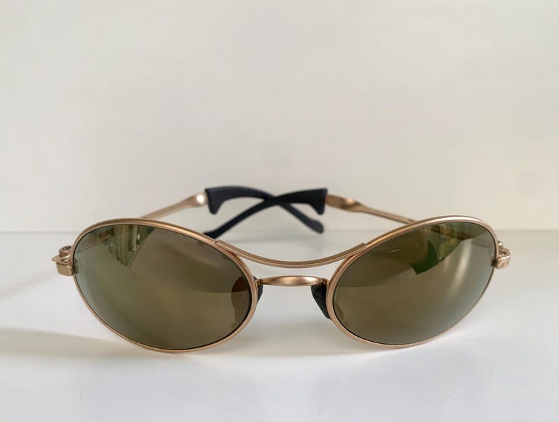 May include: Oval-shaped sunglasses with a gold-colored frame and dark lenses. The sunglasses have a double bridge design and black temples. The lenses reflect the surrounding environment.