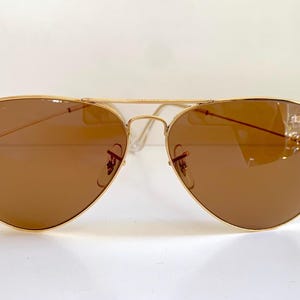 May include: Gold-framed aviator sunglasses with brown lenses. The classic teardrop-shaped lenses are set in a thin, gold metal frame. The temples and bridge are also gold, creating a sleek, timeless look. The sunglasses are designed to provide sun protection.