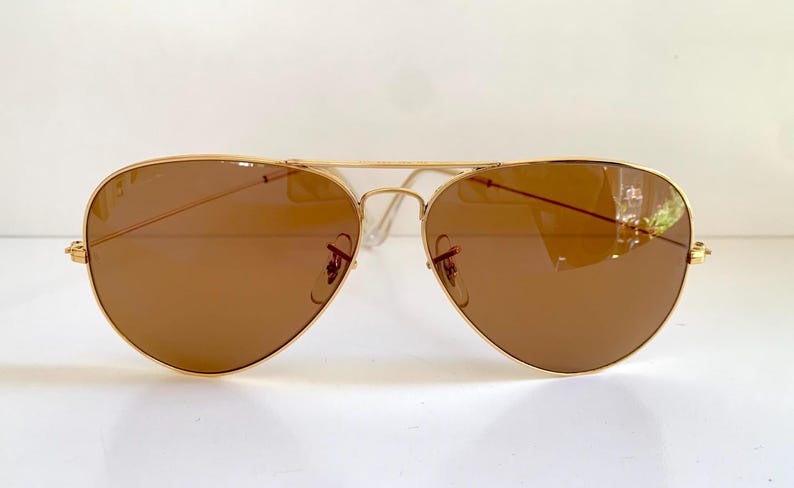 May include: Gold-framed aviator sunglasses with brown lenses. The classic teardrop-shaped lenses are set in a thin, gold metal frame. The temples and bridge are also gold, creating a sleek, timeless look. The sunglasses are designed to provide sun protection.