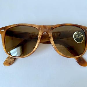 Puede incluir: Un par de gafas de sol Ray-Ban de carey marr&oacute;n con lentes marrones. Las gafas de sol tienen una barra de metal sobre el puente de la nariz y un logotipo de Ray-Ban en la lente derecha.