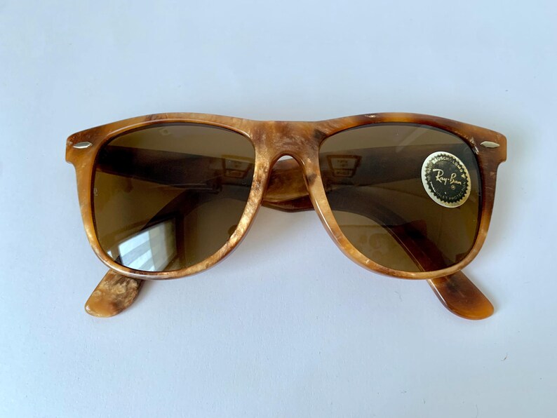 Puede incluir: Un par de gafas de sol Ray-Ban de carey marr&oacute;n con lentes marrones. Las gafas de sol tienen una barra de metal sobre el puente de la nariz y un logotipo de Ray-Ban en la lente derecha.