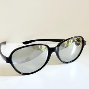May include: Black sunglasses with silver mirrored lenses. The sunglasses have a rounded frame shape and a small logo on the right lens. The sunglasses are on a white surface.