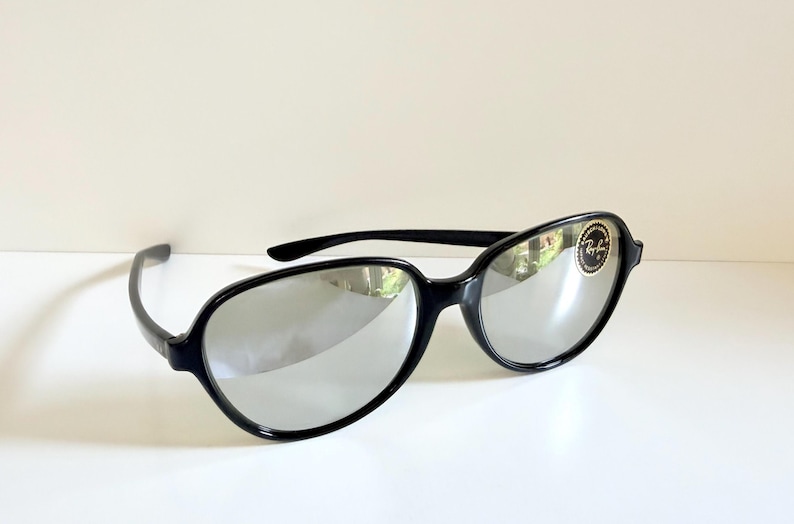 May include: Black sunglasses with silver mirrored lenses. The sunglasses have a rounded frame shape and a small logo on the right lens. The sunglasses are on a white surface.