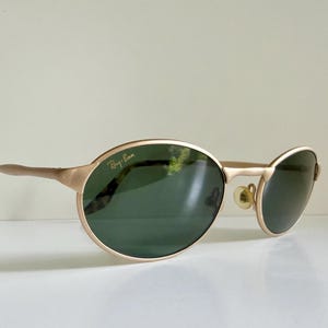 Puede incluir: Gafas de sol Ray-Ban doradas con montura ovalada y lentes verde oscuro. Las patillas tienen un patr&oacute;n de carey cerca de la oreja. El logotipo de Ray-Ban es visible en la lente superior izquierda.