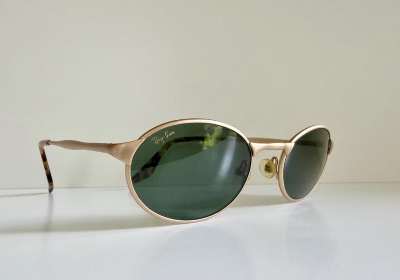 Puede incluir: Gafas de sol Ray-Ban doradas con montura ovalada y lentes verde oscuro. Las patillas tienen un patr&oacute;n de carey cerca de la oreja. El logotipo de Ray-Ban es visible en la lente superior izquierda.
