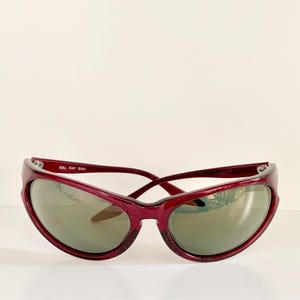 May include: A pair of vintage Ray-Ban sunglasses with a deep red frame and gray lenses. The sunglasses have a curved, sporty design. The text "B&L RAY BAN" is printed on the left arm of the sunglasses.