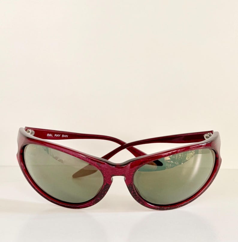 May include: A pair of vintage Ray-Ban sunglasses with a deep red frame and gray lenses. The sunglasses have a curved, sporty design. The text "B&L RAY BAN" is printed on the left arm of the sunglasses.