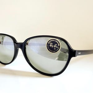 May include: Black Ray-Ban sunglasses with large, rounded frames and reflective lenses. The sunglasses have a sticker with the Ray-Ban logo. The arms of the sunglasses have two small white dots.