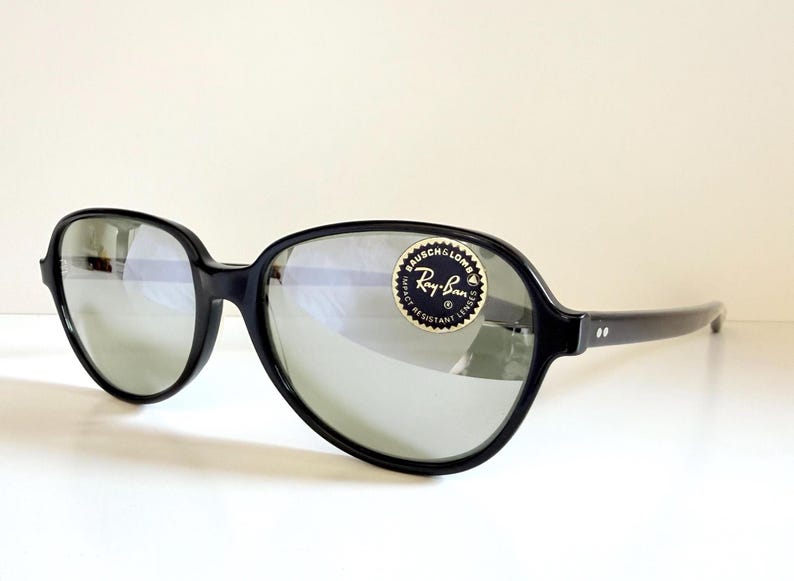 May include: Black Ray-Ban sunglasses with large, rounded frames and reflective lenses. The sunglasses have a sticker with the Ray-Ban logo. The arms of the sunglasses have two small white dots.