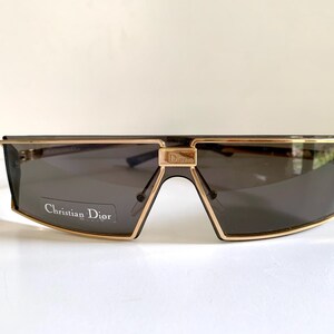 Peut inclure: Lunettes de soleil Christian Dior avec une monture dor&eacute;e et des verres gris fonc&eacute;. Les lunettes ont une forme rectangulaire avec un pont distinctif et le nom de la marque est visible sur la lentille.