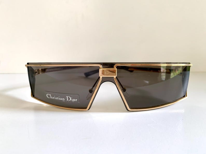 Peut inclure: Lunettes de soleil Christian Dior avec une monture dor&eacute;e et des verres gris fonc&eacute;. Les lunettes ont une forme rectangulaire avec un pont distinctif et le nom de la marque est visible sur la lentille.