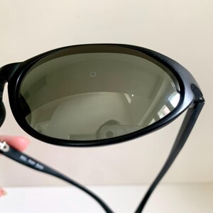 May include: Black sunglasses featuring oval-shaped dark tinted lenses with a small circular logo detail on the surface. The frames are constructed from a matte black material, and the inner temple arm displays white printed text and markings.