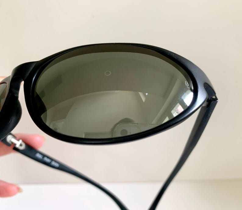 May include: Black sunglasses featuring oval-shaped dark tinted lenses with a small circular logo detail on the surface. The frames are constructed from a matte black material, and the inner temple arm displays white printed text and markings.