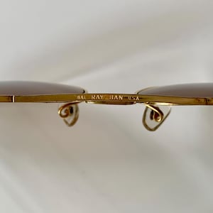 May include: Close-up of gold-rimmed sunglasses with brown lenses. The text "B&L RAY-BAN USA" is printed on the bridge. The temples have a curved design. The sunglasses are set against a white background.