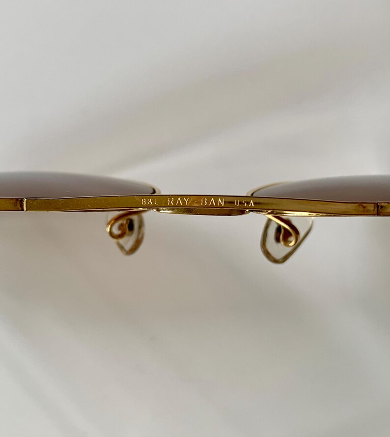 May include: Close-up of gold-rimmed sunglasses with brown lenses. The text "B&L RAY-BAN USA" is printed on the bridge. The temples have a curved design. The sunglasses are set against a white background.