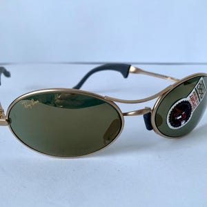 May include: Gold-tone sunglasses with oval lenses and a distinctive frame design. The lenses are a dark green color, and the temples have black accents. A sticker is on the right lens.
