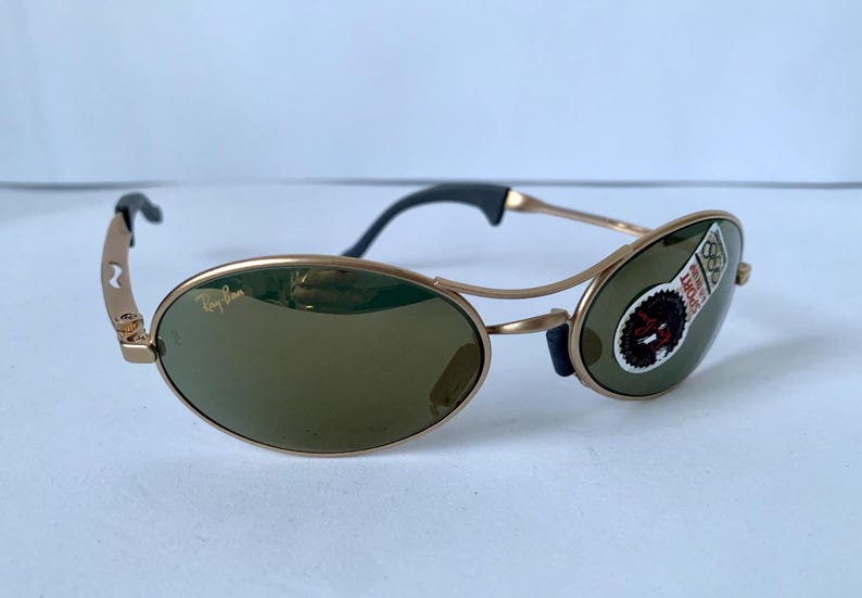 May include: Gold-tone sunglasses with oval lenses and a distinctive frame design. The lenses are a dark green color, and the temples have black accents. A sticker is on the right lens.