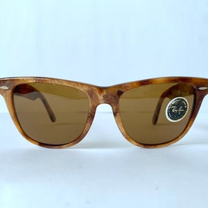 Puede incluir: Gafas de sol Ray-Ban de carey marr&oacute;n con lentes marrones. Las gafas tienen una barra de metal en la parte superior del marco y una pegatina del logotipo de Ray-Ban en la lente derecha.
