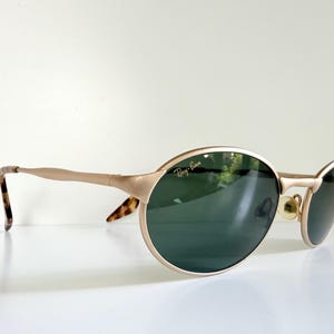 Puede incluir: Gafas de sol Ray-Ban doradas con lentes ovaladas y una sutil barra en la ceja. Las lentes son de color verde oscuro y las patillas tienen un patr&oacute;n de carey en los extremos. El logotipo de Ray-Ban es visible en la lente izquierda.