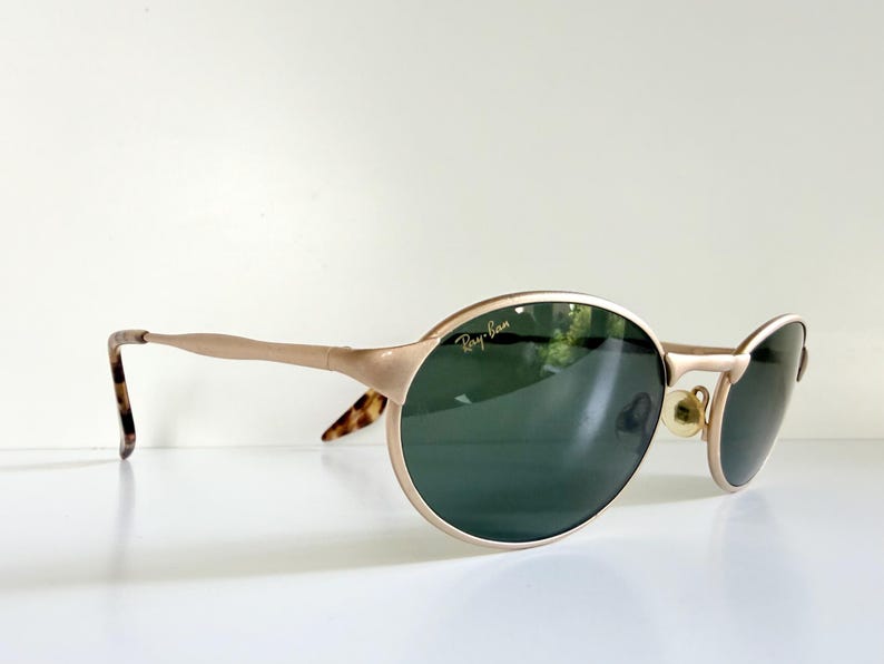 Puede incluir: Gafas de sol Ray-Ban doradas con lentes ovaladas y una sutil barra en la ceja. Las lentes son de color verde oscuro y las patillas tienen un patr&oacute;n de carey en los extremos. El logotipo de Ray-Ban es visible en la lente izquierda.