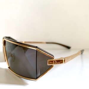Peut inclure: Lunettes de soleil g&eacute;om&eacute;triques avec une monture dor&eacute;e et des verres fonc&eacute;s. Les lunettes pr&eacute;sentent un design angulaire distinctif avec le nom de la marque "Dior" affich&eacute; sur la branche. Les branches sont de couleur or.