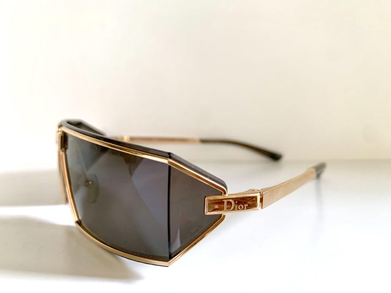Peut inclure: Lunettes de soleil g&eacute;om&eacute;triques avec une monture dor&eacute;e et des verres fonc&eacute;s. Les lunettes pr&eacute;sentent un design angulaire distinctif avec le nom de la marque "Dior" affich&eacute; sur la branche. Les branches sont de couleur or.