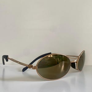 May include: Gold-tone sunglasses with oval lenses. The frames are a light gold color, with dark blue accents on the temples. The lenses are a brown tint, and the sunglasses are resting on a white surface.