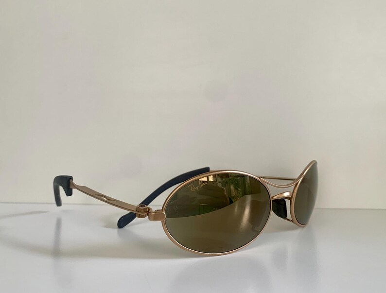 May include: Gold-tone sunglasses with oval lenses. The frames are a light gold color, with dark blue accents on the temples. The lenses are a brown tint, and the sunglasses are resting on a white surface.