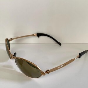 May include: Gold-toned sunglasses with oval lenses and black temple tips. The frames have a curved design with a subtle wave detail on the temples. The lenses are a dark, neutral color, providing a sleek, modern look.