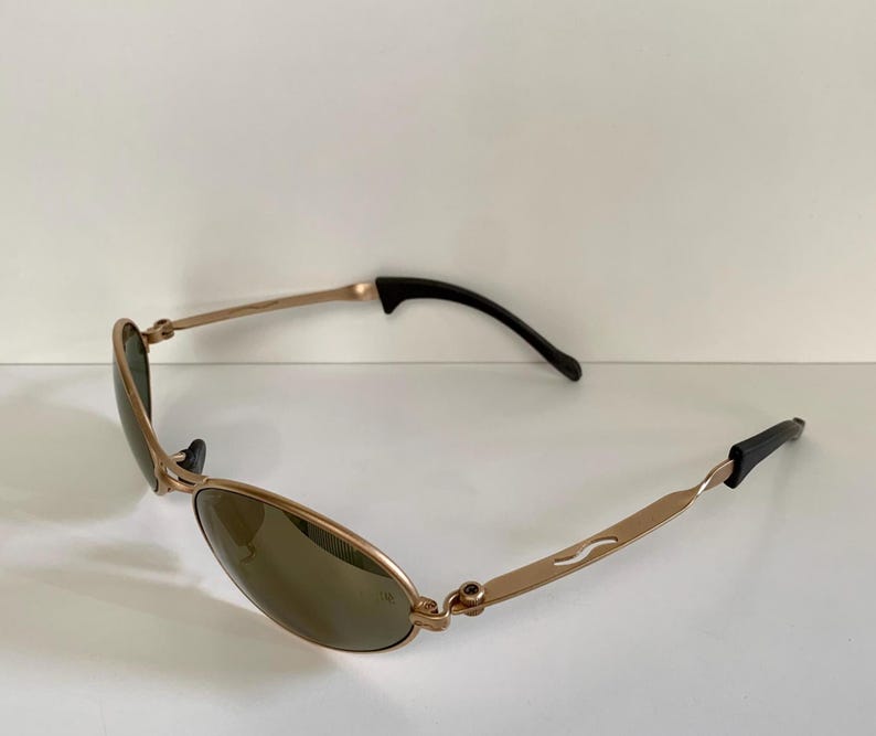 May include: Gold-toned sunglasses with oval lenses and black temple tips. The frames have a curved design with a subtle wave detail on the temples. The lenses are a dark, neutral color, providing a sleek, modern look.