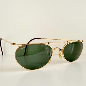 K&ouml;nnte beinhalten: Goldfarbene Sonnenbrille mit gr&uuml;nen Gl&auml;sern. Die Sonnenbrille hat einen Doppelsteg und einen klassischen Aviator-Stil. Die B&uuml;gel sind dunkelbraun. Das Ray-Ban-Logo ist auf dem Glas sichtbar.