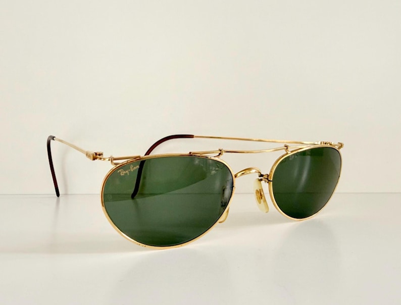 K&ouml;nnte beinhalten: Goldfarbene Sonnenbrille mit gr&uuml;nen Gl&auml;sern. Die Sonnenbrille hat einen Doppelsteg und einen klassischen Aviator-Stil. Die B&uuml;gel sind dunkelbraun. Das Ray-Ban-Logo ist auf dem Glas sichtbar.