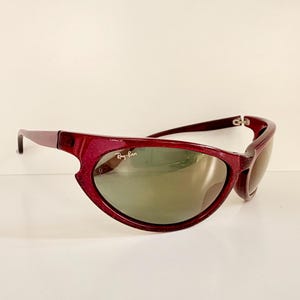 May include: Maroon-colored Ray-Ban sunglasses with a curved frame and green-tinted lenses. The Ray-Ban logo is visible on the lens. The sunglasses have a sporty, wraparound design.