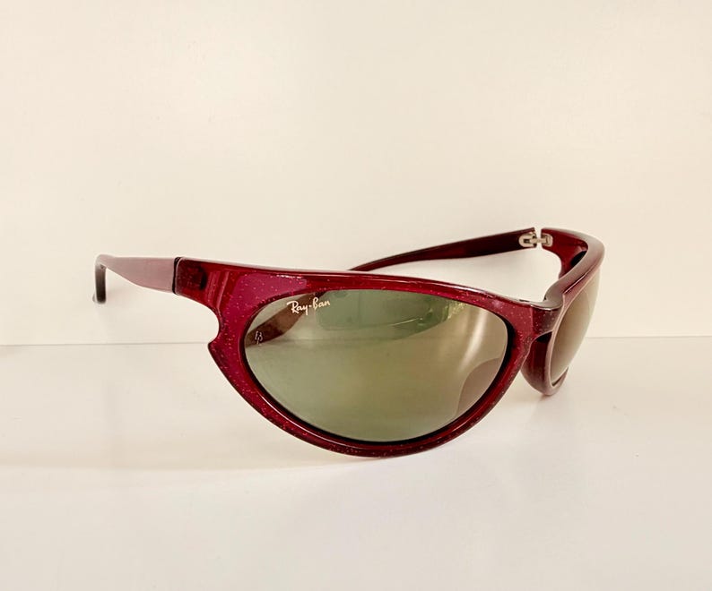 May include: Maroon-colored Ray-Ban sunglasses with a curved frame and green-tinted lenses. The Ray-Ban logo is visible on the lens. The sunglasses have a sporty, wraparound design.