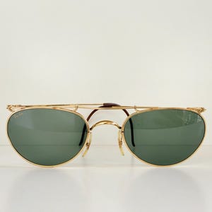 K&ouml;nnte beinhalten: Goldgerahmte Sonnenbrille mit gr&uuml;nen Gl&auml;sern. Die Sonnenbrille hat einen Doppelsteg und eine markante Form. Die Gl&auml;ser sind oval und der Rahmen ist goldfarben. Auf einem schlichten wei&szlig;en Hintergrund.