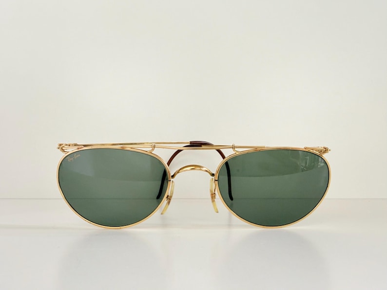 K&ouml;nnte beinhalten: Goldgerahmte Sonnenbrille mit gr&uuml;nen Gl&auml;sern. Die Sonnenbrille hat einen Doppelsteg und eine markante Form. Die Gl&auml;ser sind oval und der Rahmen ist goldfarben. Auf einem schlichten wei&szlig;en Hintergrund.